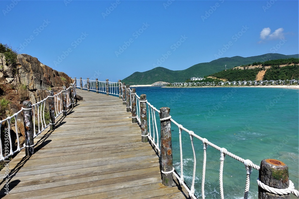 Summer. Sunny day. A wooden path with rope handrails goes over the turquoise sea around the mountain into the unknown. In the distance are green hills, a white beach and small houses.