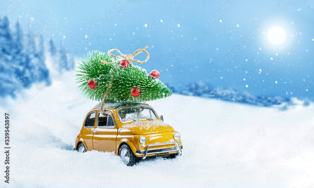 Retro toy car carrying christmas tree on roof in snowy winter forest. Christmas background. Holidays card. Copy space.