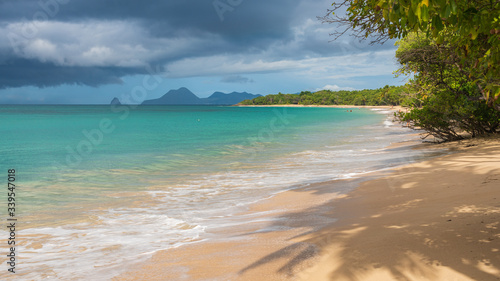 Écume d'une vague sur une plage de sable blanc avec la mer bleu turquoise, un ciel ensoleillé avec des nuages noirs menaçants, la mangrove à l'arrière plan et les reliefs de la martinique à la Saline