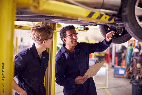 Male Tutor With Student Looking Underneath Car On Hydraulic Ramp On Auto Mechanic Course  © Monkey Business