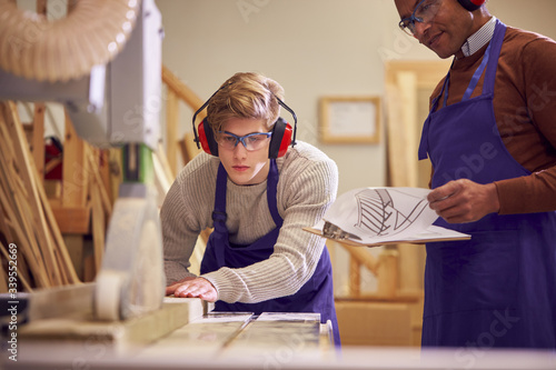 Tutor With Male Carpentry Student In Workshop Studying For Apprenticeship At College Using Bench Saw