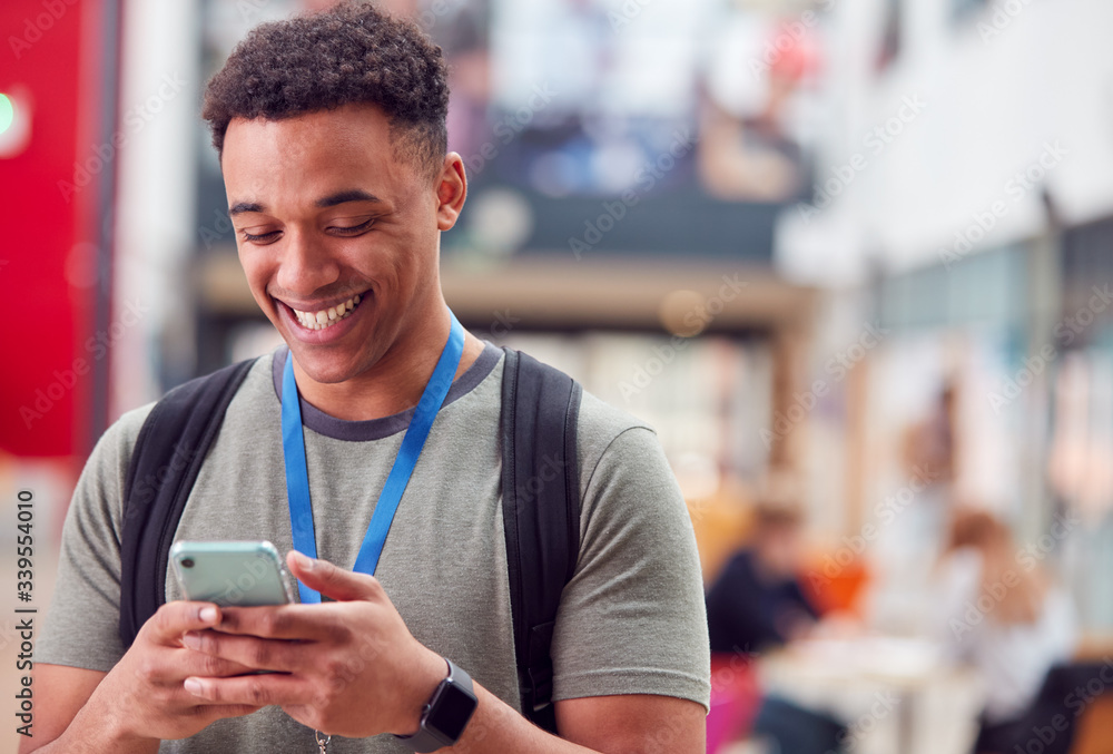 Smiling Male College Student Checking Mobile Phone In Busy Communal Campus Building Smiling Male College Student Checking Mobile Phone In Busy Communal Campus Building
