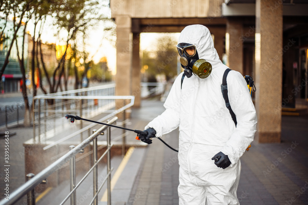 Man sprays disinfector onto the railing wearing coronavirus protective suit and equipment. Cleaning and sterilizing the not crowded city streets. Covid-19 nCov2019 spread prevention.