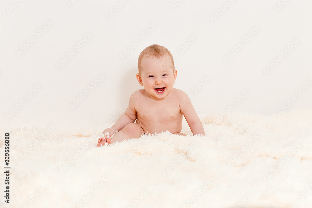 a naked Caucasian eight-month-old baby sits on a bed against a white background and laughs. a naked Caucasian eight-month-old baby sits on a bed against a white background and laughs.