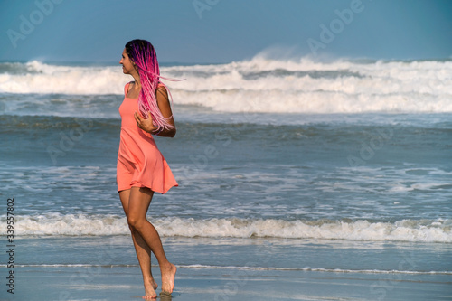 Beautiful bright girl in a pink dress with long pink dreadlocks on the beach. Pacific ocean, tide, storm, big waves