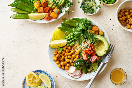 Healthy vegetarian lunch bowl with avocado, chickpeas, quinoa and vegetables, garnished with microgreens and nut dressing. Flat lay on stone background.