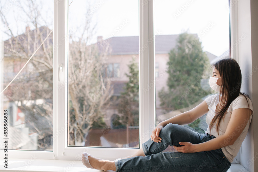 Side view of Young woman sits on windowsill and in protective mask and look out the window. Alone at home in quarantine. Coronavirus theme. Stay at home Side view of Young woman sits on windowsill and in protective mask and look out the window. Alone at home in quarantine. Coronavirus theme. Stay at home