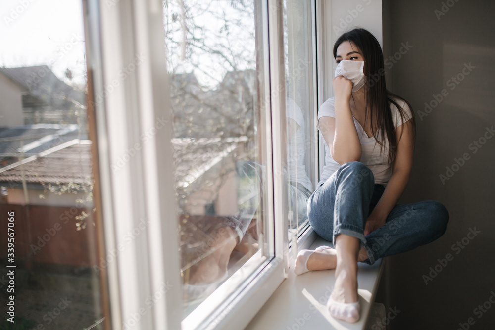 Side view of Young woman sits on windowsill and in protective mask and look out the window. Alone at home in quarantine. Coronavirus theme. Stay at home Side view of Young woman sits on windowsill and in protective mask and look out the window. Alone at home in quarantine. Coronavirus theme. Stay at home