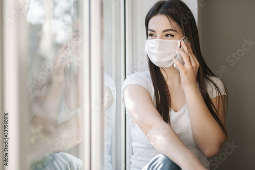 Attractive woman in medical mask sits on windowsill at home. Female in white t'shirt and white medical mask use phone