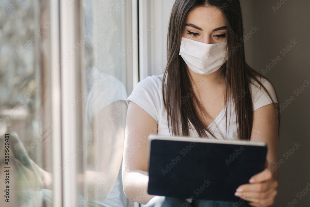 Attractive young woman in white medical mask sits on windowsiil and use tablet. Female have a rest at home in quarantine Attractive young woman in white medical mask sits on windowsiil and use tablet. Female have a rest at home in quarantine