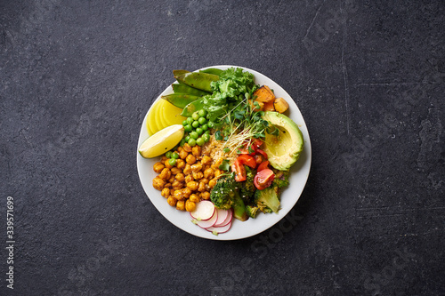 Flat lay with healthy vegetarian lunch bowl with avocado, chickpeas, quinoa and vegetables, garnished with microgreens and nut dressing. Dark concrete background, copy space.