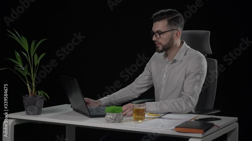 Young attractive businessman with beard in shirt puts on glasses and begins work at laptop. Studio shot with black background.