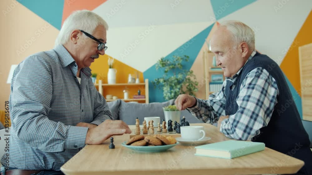 Cheerful elderly men are playing chess at home talking sitting at table together enjoying intelligent sports. Entertainment and friendship concept. Cheerful elderly men are playing chess at home talking sitting at table together enjoying intelligent sports. Entertainment and friendship concept.