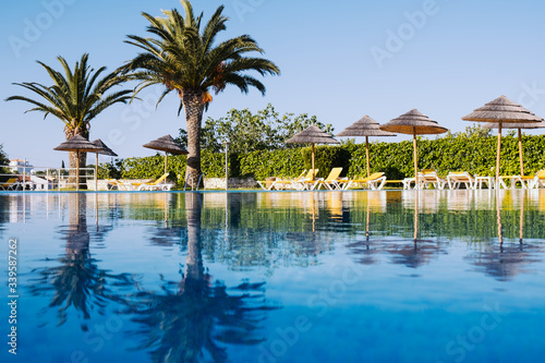 Beautiful tropical beach front hotel resort with swimming pool, sun-loungers and palm trees during a warm sunny day.