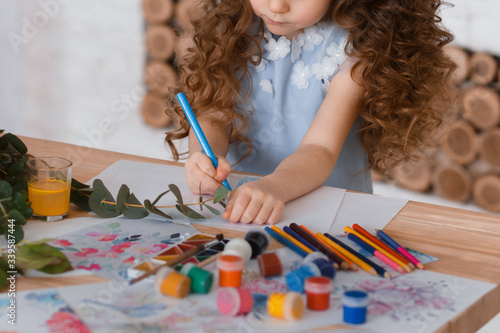 Girl with curly hair sits at a table draws with a blue pencil. She is dressed in a blue dress with flowers. On the table are pencils, flowers, ribbons. Mother's day Selective focus. Medium sharp photo