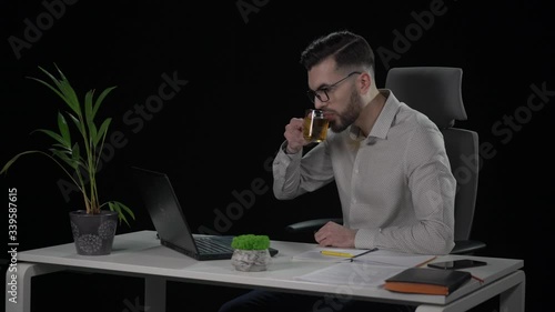 Caucasian attractive bearded male copywriter in glasses typing text from paper on laptop and drinking tea at white desk. Indoor studio shot on black background.