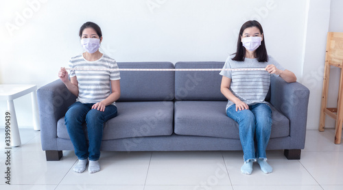 Foto Asian women wearing masks sit on sofa spaced apart and hold measuring tape for social distancing to reduce spreading of coronavirus