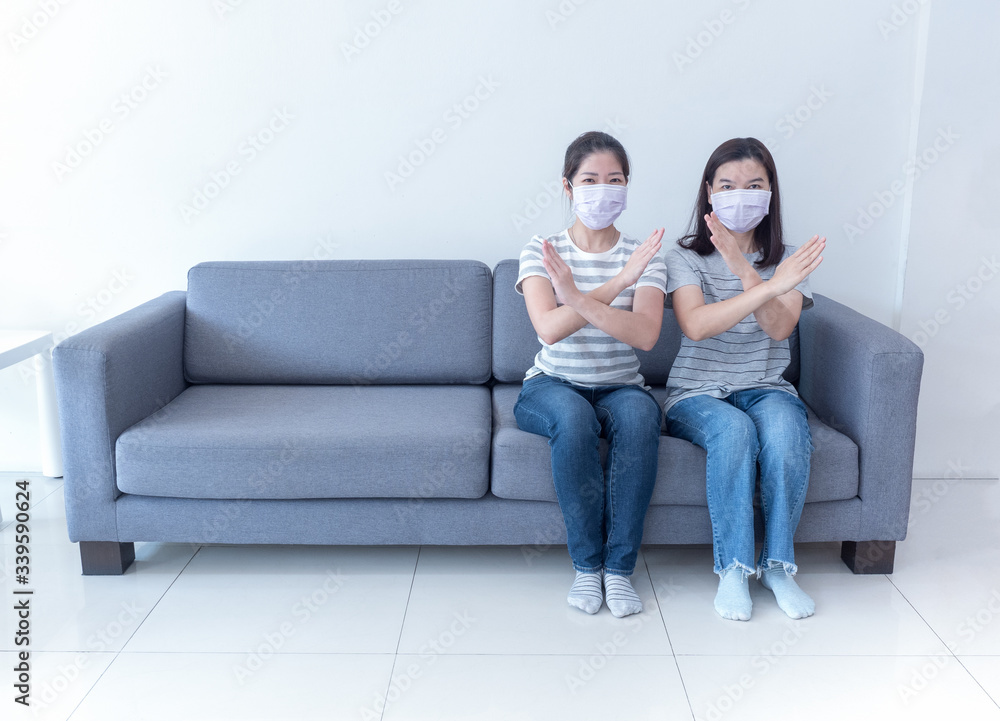 Asian women wearing masks sit together on sofa without space and to cross sign for social distancing to reduce spreading of coronavirus. Work from home, social distancing and covid-19 concept. Asian women wearing masks sit together on sofa without space and to cross sign for social distancing to reduce spreading of coronavirus. Work from home, social distancing and covid-19 concept.