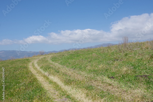 Panoramic view of a highland meadow under blue sky with some white clouds and a mountain ridge in the distant background on a spring day