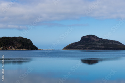 Two small islands or rocky landmasses, blue sky with clouds and they are surrounded by a calm deep blue ocean. The land is reflecting in the water. The islands are uninhabited small tropical 