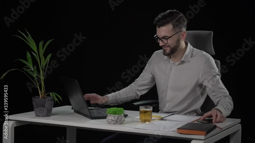 Caucasian attractive bearded man in glasses working on laptop and reciving pleasant message on his smartphone at white desk. Indoor studio shot on black background.
