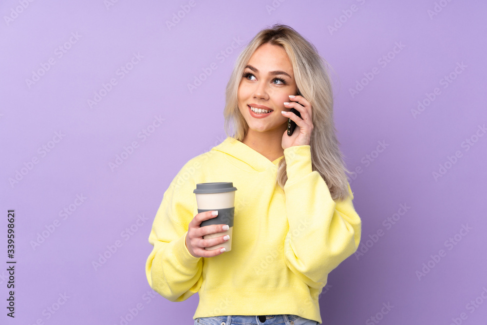 Teenager girl wearing a yellow sweatshirt over isolated purple background holding coffee to take away and a mobile Teenager girl wearing a yellow sweatshirt over isolated purple background holding coffee to take away and a mobile