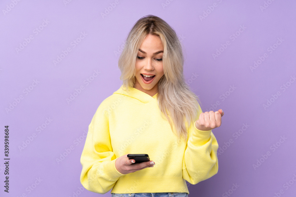 Teenager girl wearing a yellow sweatshirt over isolated purple background surprised and sending a message Teenager girl wearing a yellow sweatshirt over isolated purple background surprised and sending a message