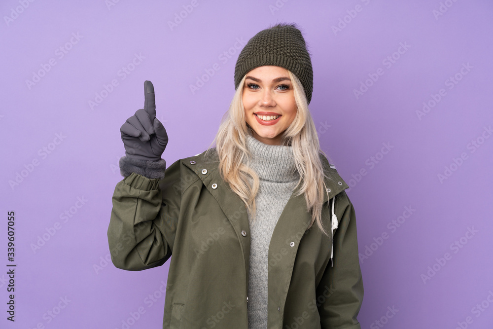 Teenager blonde girl with winter hat over isolated purple background showing and lifting a finger in sign of the best Teenager blonde girl with winter hat over isolated purple background showing and lifting a finger in sign of the best