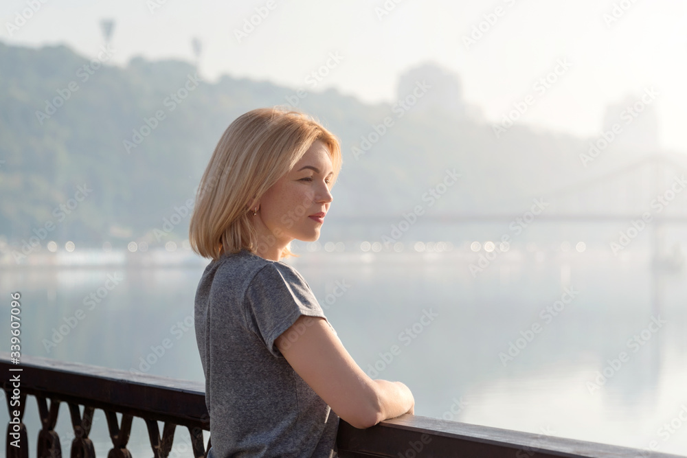 Young blonde woman enjoying city view from the bridge. Lovely woman spending time on the river embankment at sunset. Soft backlit. Lifestyle relax recreation weekend concept