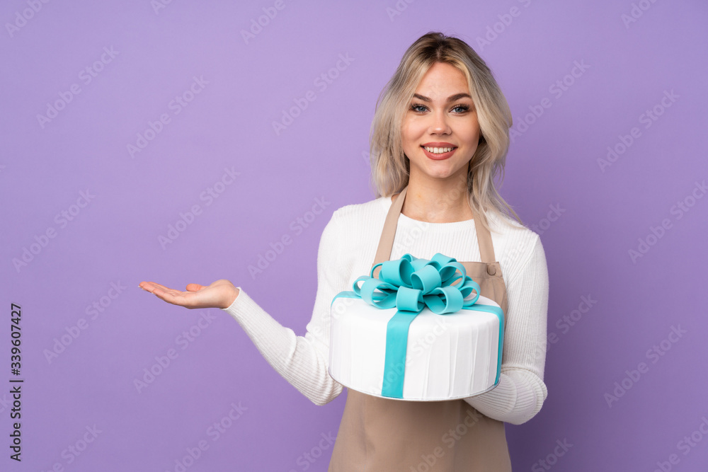 Young pastry chef holding a big cake over isolated purple background holding copyspace imaginary on the palm