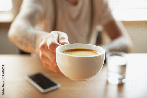 Close up of male hands proposing cup of coffee, sitting at the table with smartphone. Surfing, online shopping, working. Education, freelance, art and business concept. Drinking. Hot aroma drink.