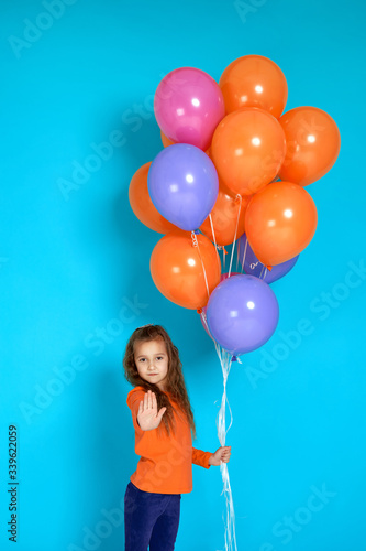 little child girl with bright colorful air balloons showing stop gesture isolated on blue background. birthday party.