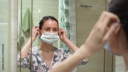 Portrait of attractive Caucasian brunette woman in front of the mirror in bathroom. Girl in floral night robe puts on a blue medical face mask. Female begin new day at home in quarantine isolation.