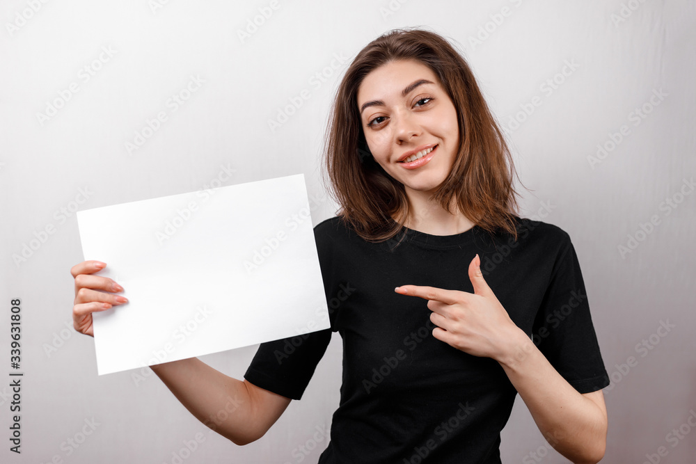 young brunette woman in a black t-shirt smiling shows a white sheet on a white background. copyspase young brunette woman in a black t-shirt smiling shows a white sheet on a white background. copyspase