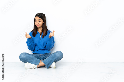 Young mixed race woman sitting on the floor isolated on white background making money gesture