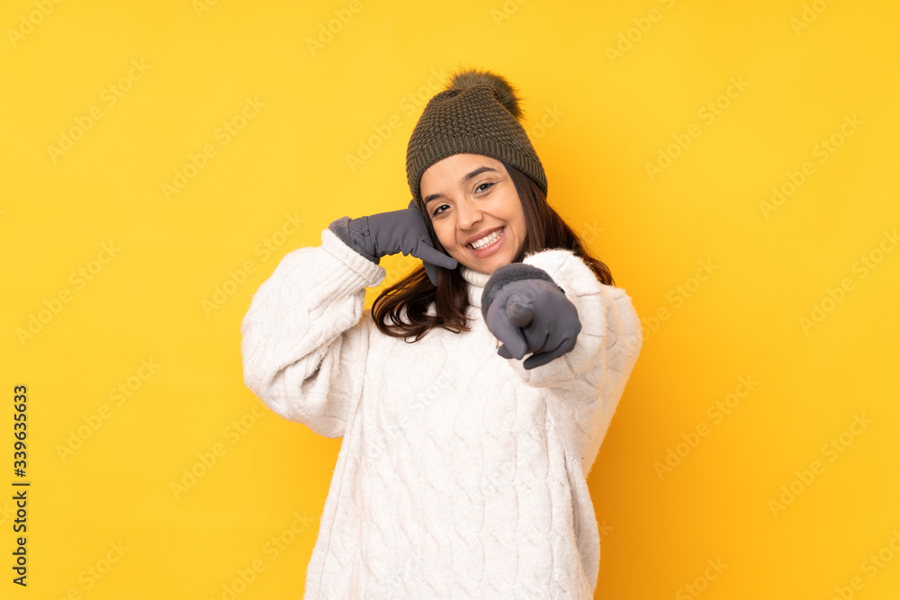 Young woman with winter hat over isolated yellow background making phone gesture and pointing front Young woman with winter hat over isolated yellow background making phone gesture and pointing front