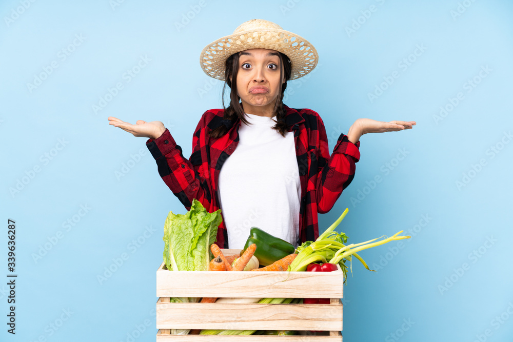 Young farmer Woman holding fresh vegetables in a wooden basket having doubts while raising hands