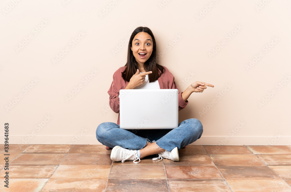 Young mixed race woman with a laptop sitting on the floor surprised and pointing side