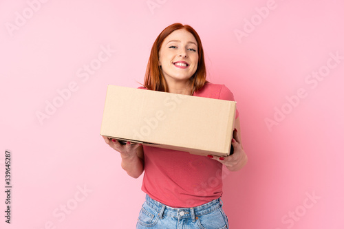 Young redhead woman over isolated pink background holding a box to move it to another site