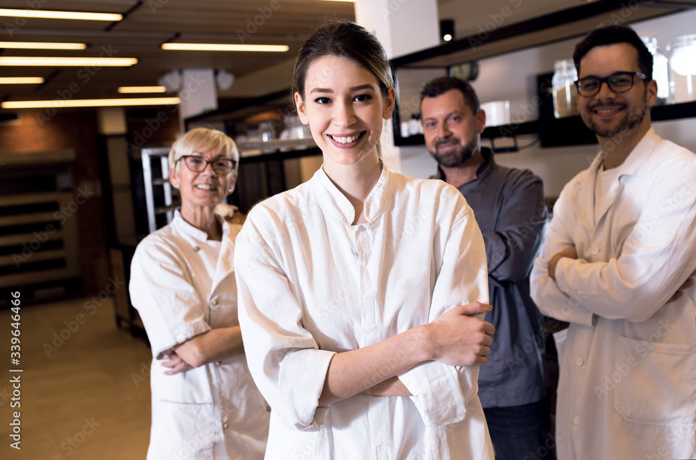 Portrait of young female baker at bakery with her colleagues in backgrounds. Portrait of young female baker at bakery with her colleagues in backgrounds.