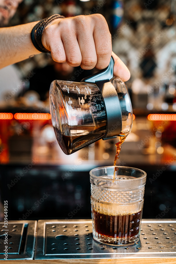 Vertical image. Alternative Coffee Brewing Method. Close-up of the hands of barista, pouring a black filter coffee from a teapot into a transparent glass on a bar counter in a cafe.