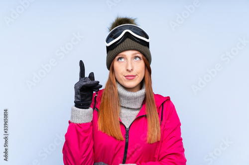 Skier redhead woman with snowboarding glasses over isolated blue wall pointing with the index finger a great idea