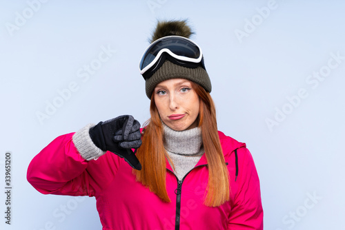 Skier redhead woman with snowboarding glasses over isolated blue wall showing thumb down sign