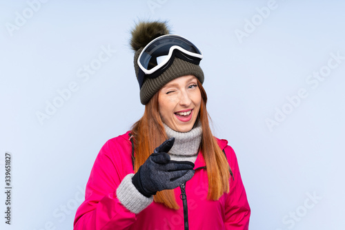 Skier redhead woman with snowboarding glasses over isolated blue wall points finger at you