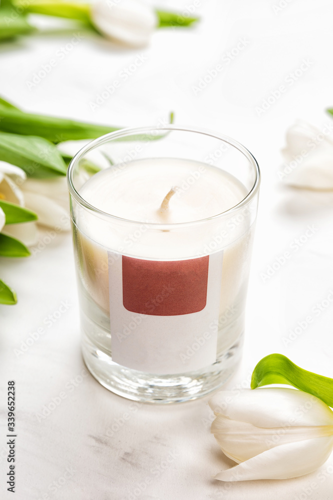 Aroma candle in glass jar and tulip flowers on white marble background, front view. Aromatherapy spa concept
