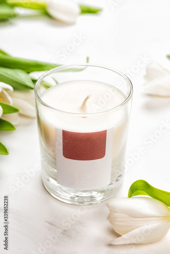 Aroma candle in glass jar and tulip flowers on white marble background, front view. Aromatherapy spa concept