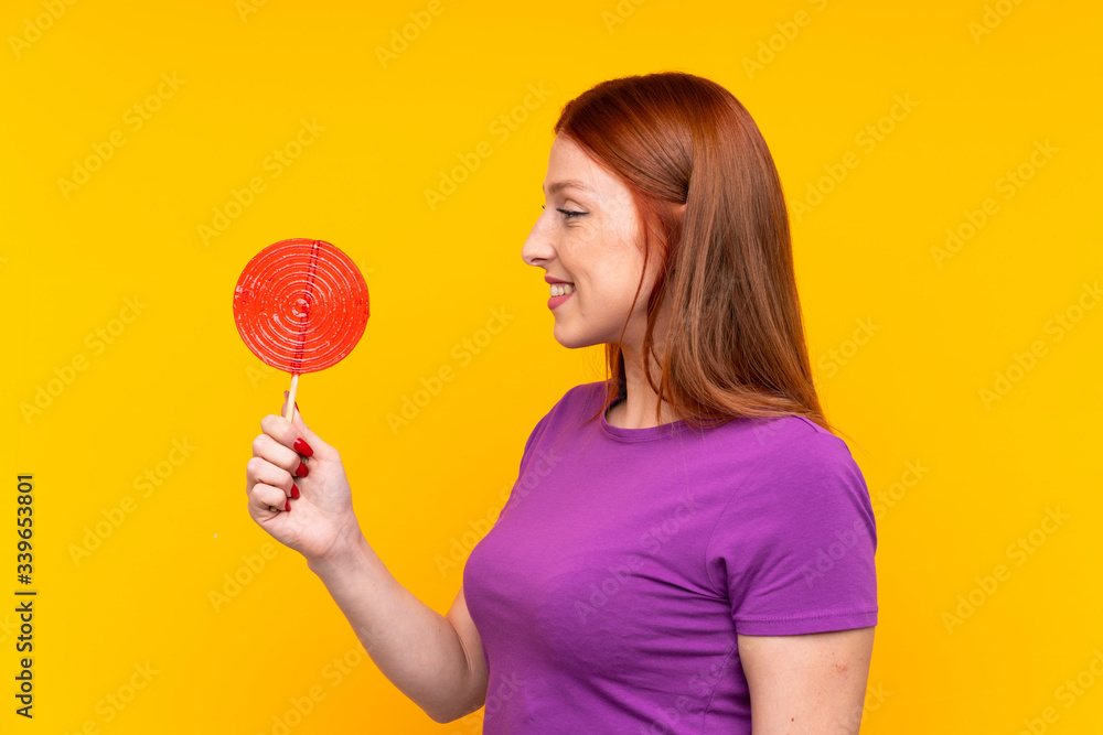 Young redhead woman holding a lollipop over yellow background with happy expression Young redhead woman holding a lollipop over yellow background with happy expression