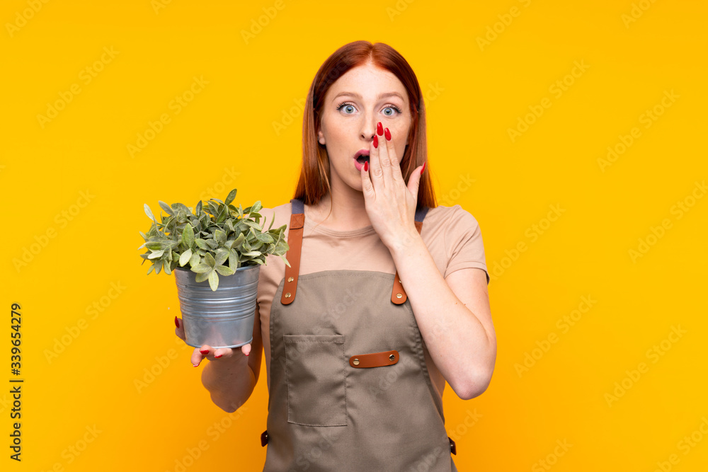 Young redhead gardener woman holding a plant over isolated yellow background with surprise facial expression Young redhead gardener woman holding a plant over isolated yellow background with surprise facial expression