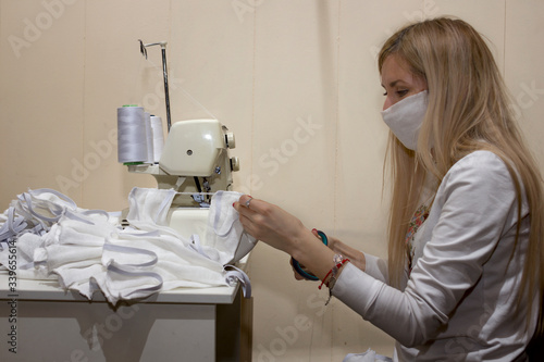 blonde girl with a white mask on her face sews gauze protective masks on an overlock sewing machine to protect against coronavirus during an epidemic photo