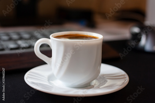 white espresso cup on a saucer in front of a keyboard in the workplace. Hot coffee while working day. Cozy calm atmosphere at the workplace. Fragrant refreshing drink in mug. Coffee break concept